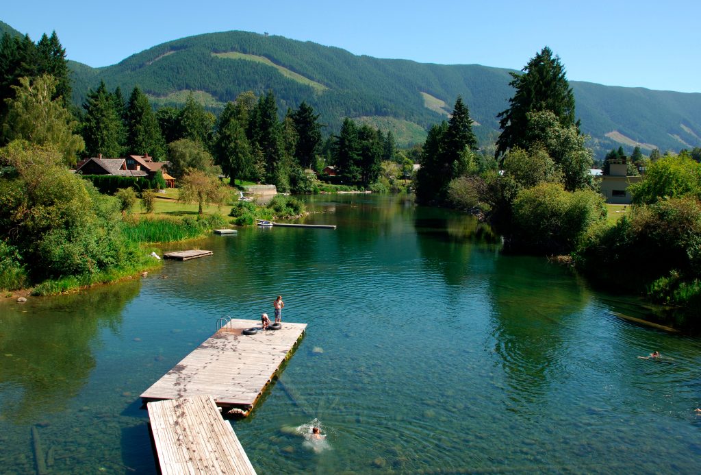 People swimming from a dock in the clear water near Lake Cowichan, surrounded by forested hills and waterfront homes.