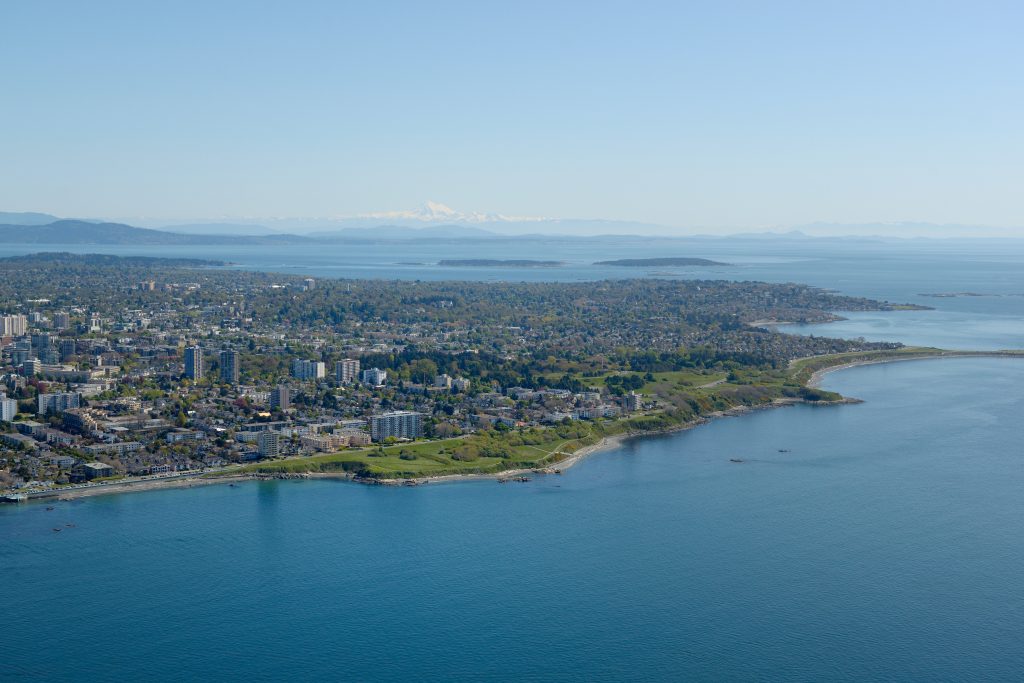Aerial view of Oak Bay and the surrounding Greater Victoria coastline, with ocean, residential neighbourhoods, green parkland, and distant mountains in the background.